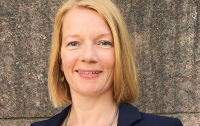 Professional headshot of a smiling blonde woman wearing a dark blazer, standing against a stone wall